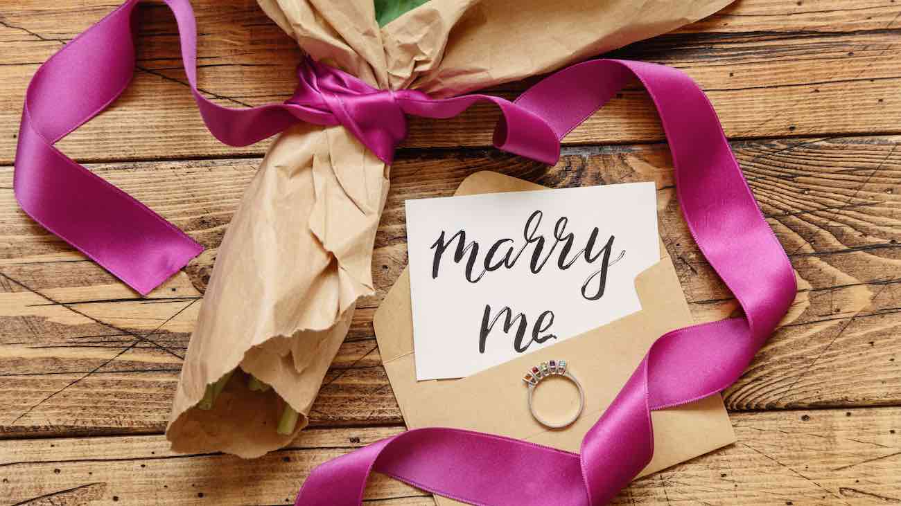 Bouquet in craft paper with a MARRY ME card and proposal ring on wooden table